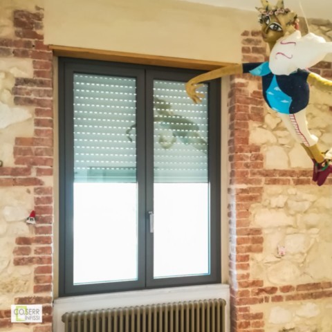 Finestra a due ante in allumnio installata in appartamento add Augusta da Coserr Serramento a due ante in alluminio scuro su muro di pietra chiara con pupazzo in cartapesta che suona il violino in appartamento ad Augusta