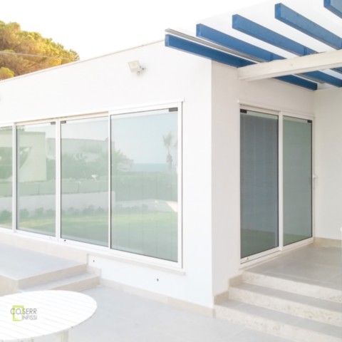 Porte finestre scorrevoli con veneziane Pellini interne al vetrocamera per villa al mare a Siracusa Facciata di villetta al mare con infissi scorrevoli bianche con veneziane Pellini interne al vetrocamera e pegolato azzurro a Siracusa